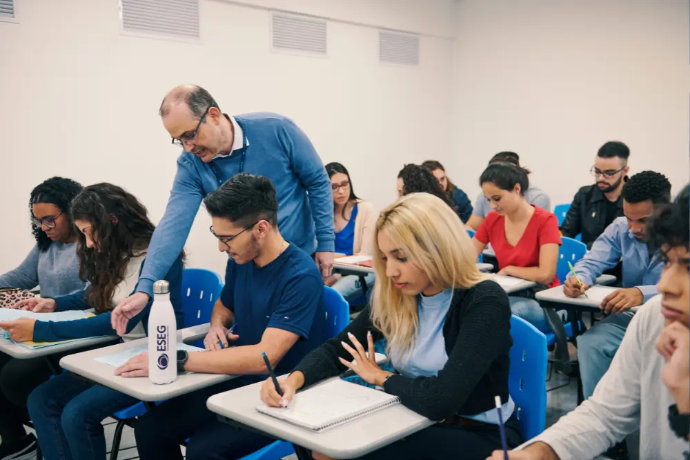 Professor auxiliando alunos durante uma aula na Faculdade ESEG, com estudantes focados em suas anotações em um ambiente de sala de aula moderna, com garrafa personalizada da faculdade ESEG sobre a mesa.