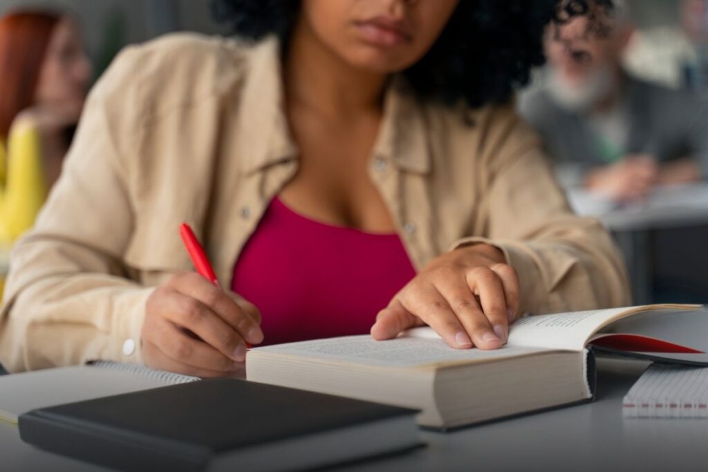Estudante realizando leitura e anotações em um livro aberto, sentada em ambiente acadêmico, com cadernos e materiais de estudo sobre a mesa.