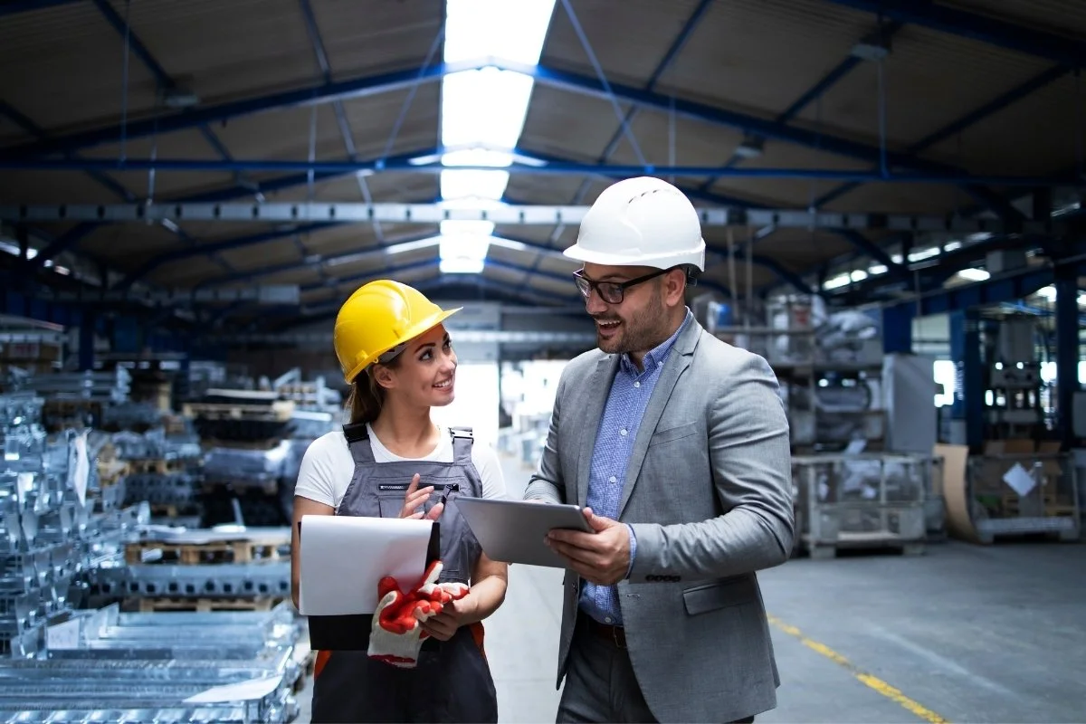 Dois profissionais de Engenharia de Produção usando capacetes de segurança analisam um tablet dentro de uma fábrica, cercados por equipamentos industriais e linhas de produção.