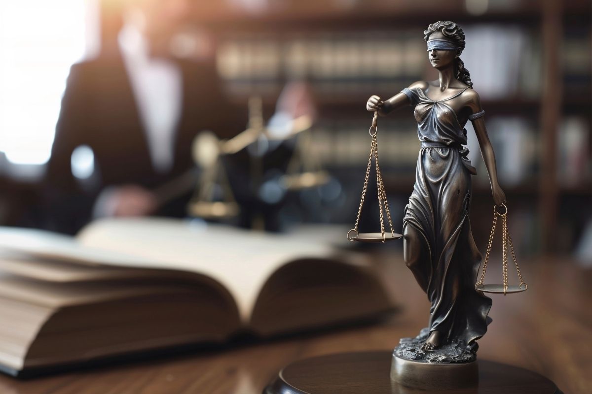 Estátua da Justiça com os olhos vendados e uma balança nas mãos, diante de um livro aberto e de um profissional do Direito ao fundo.