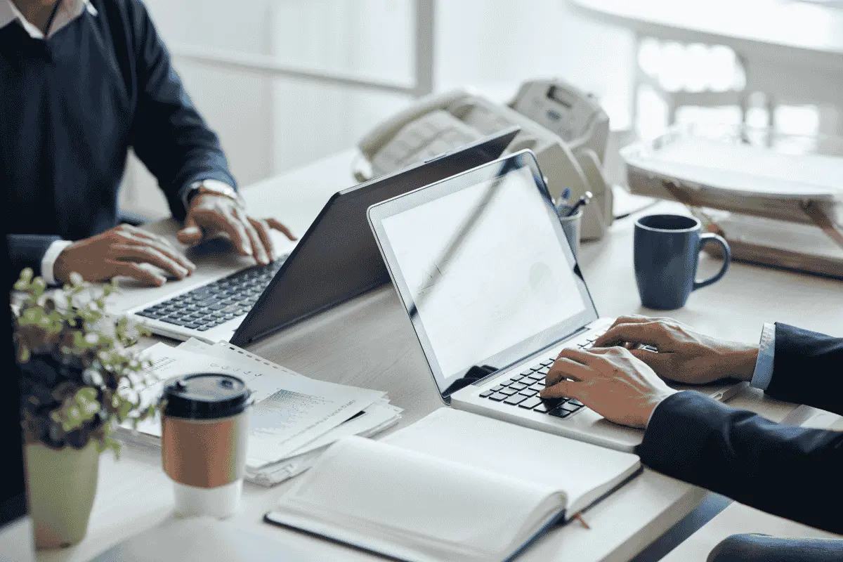 Pessoa de terno vista de costas usando um laptop com gráficos em uma reunião híbrida de trabalho, com colegas à mesa e um apresentador em uma tela ao fundo.